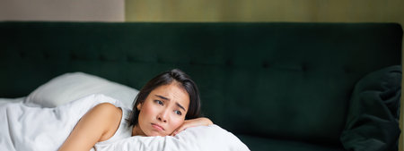 Sad and gloomy asian girl lying in her bed, grimacing and crying, feeling upset, spending time in...