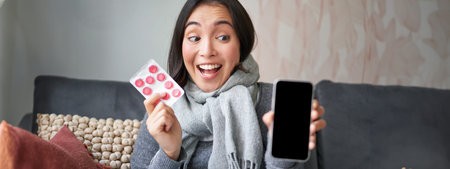 Happy asian woman catching cold, showing smartphone screen and pills, recommending online doctor...
