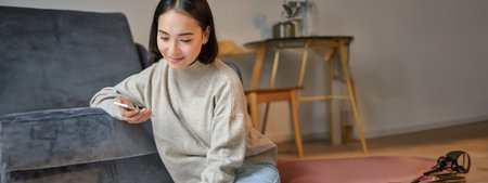 Vertical shot of young woman in cozy home working on laptop, using smartphone and drinking coffee, sitting on floor near sofaの写真素材