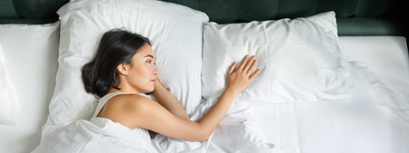 Portrait of romantic asian woman lying alone in her bed, touching empty pillow and thinking about...