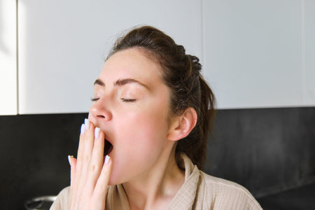 Close up portrait of tired young woman in bathrobe, yawning in the morning, making herself cup of...