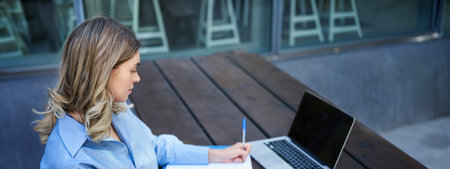 Portrait of businesswoman taking notes, studying on her computer, sitting outside in park. Young...