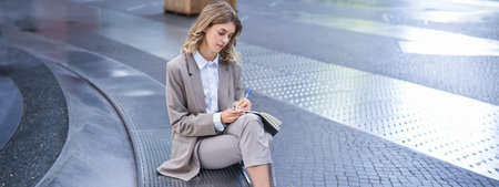 Business woman sits in city centre and works on mobile phone, she has her laptop and wears working...