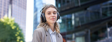 Portrait of smiling corporate woman, girl in suit listening music in headphones, sitting in city...