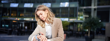 Portrait of corporate woman checks her time, stands on street outside office and looks at her wrist watchの写真素材
