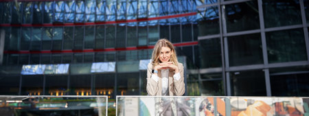 Beautiful young woman in beige suit, standing near office buildings in city center, smiling and looking dreamyの写真素材