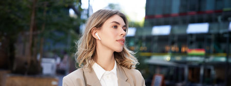 Close up portrait of blond businesswoman, confident corporate woman in headphones and beige suit, posing outdoors on street of city centerの写真素材