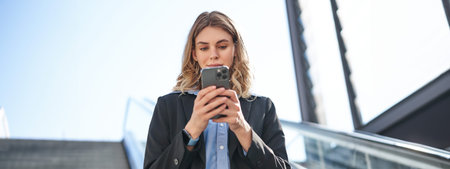 Portrait of businesswoman in suit, standing on escalator, walking in city, using mobile phone....