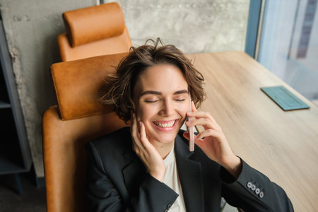 Portrait of business woman in suit, sitting in her office and answering a phone call with pleased...