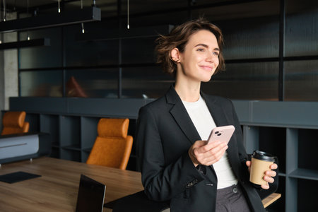 Image of young successful businesswoman in conference room, standing in an office with smartphone and drinking coffee, smiling while looking out the windowの写真素材