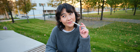Portrait of asian woman, student in park, sits on bench with digital tablet and a pen, thinking, looking aside thoughtful, making notes, does her homework outdoorsの写真素材
