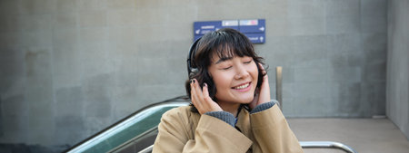Portrait of happy woman, korean girl in headphones, listening music in headphones, enjoying sound of...