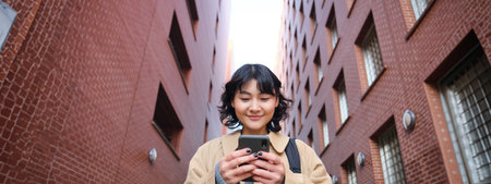 Lower angle view of brunette korean girl, listening music in headphones, walking along street and looking at smartphone, reading message on mobile phoneの写真素材