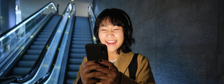 Laughing cute Korean girl on escalator, going down to tube, underground or metro, using mobile phone, watching smth funny on smartphone appの写真素材