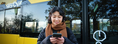 Beautiful korean girl, student on bus stop, looking at her smartphone, checking timetable, reading...