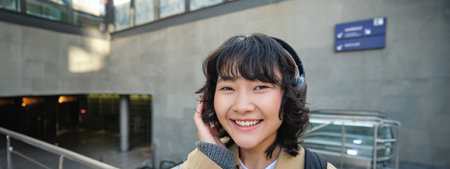 Close up of smiling brunette girl in headphones, listens music, travels around city, commutes to work, stands on a streetの写真素材