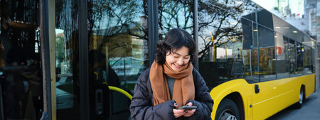 Beautiful korean girl, student on bus stop, looking at her smartphone, checking timetable, reading text message, wearing winter clothesの写真素材
