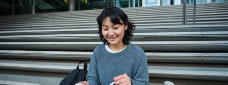 Smiling girl, graphic designer, using digital tablet and pen tool to draw, does home assignment for university, sits on stairsの写真素材