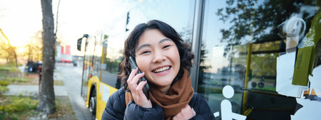 Cellular technology and people concept. Stylish asian girl talks on mobile phone, makes a telephone call, stands near bus stop and has conversationの写真素材