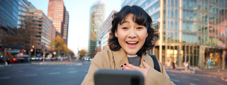Portrait of asian girl looks surprised at smartphone screen, video chats and hears amazing news, looks impressed, stands on sunny street of city centreの写真素材