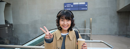 Positive korean girl in headphones, drinks coffee to go, shows peace, v-sign, stands on street and smiles happilyの写真素材