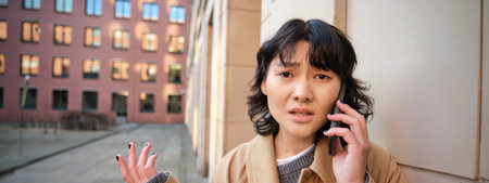 Korean girl with concerned face talks on mobile phone and shrugs, frowns and looks concerned, being lost in town, stands on streetの写真素材