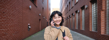 Portrait of young korean woman walking down street with phone, talking with someone, makes a call, has telephone conversationの写真素材
