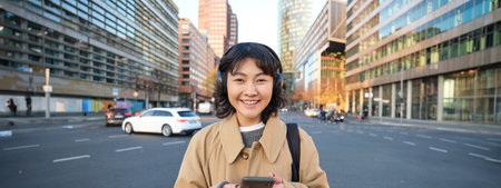 Portrait of young asian girl, student walks in city, listens music in headphones and uses mobile phone on streetsの写真素材