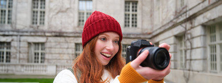Redhead girl photographer takes photos on professional camera outdoors, captures streetstyle shots, looks excited while taking picturesの写真素材