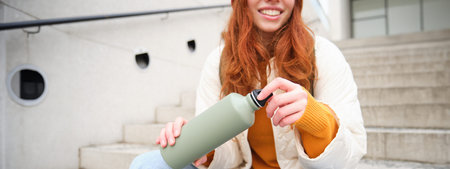 Smiling traveler, redhead girl tourist sits on stairs with flask, drinks hot coffee from a flask...