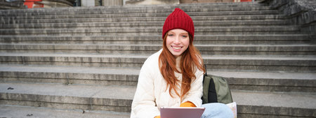 Beautiful young modern girl with red hair, holds digital tablet, sits on stairs near museum and...