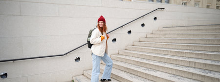 Young stylish girl traveller goes sightseeing, travels around city with backpack, tourist smiles and looks aroundの写真素材