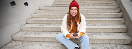 Hipster ginger girl, redhead woman sits on stairs with smartphone, waits for someone and messages on...