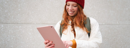 Young redhead girl, walks with digital tablet around city, stands on street with backpack and...