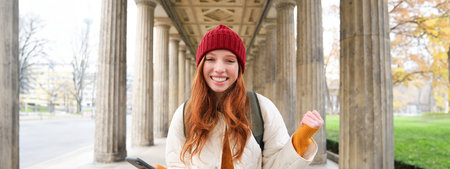 Mobile broadband and people. Smiling redhead 20s girl with backpack, uses smartphone on street,...