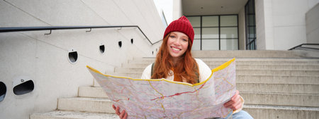 Young smiling redhead girl, tourist sits on stairs outdoors with city paper map, looking for direction, traveller backpacker explores city and looks for sightseeingの写真素材