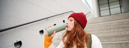 Smiling traveler, redhead girl tourist sits on stairs with flask, drinks hot coffee from a flask...