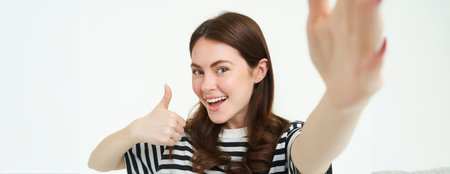 Portrait of smiling woman recommending product, shows thumbs up, takes selfie with something good, copy space, standing isolated on white backgroundの写真素材