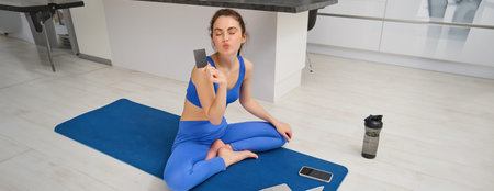 Image of smiling young woman, holding credit card, sitting on fitness mat with laptop, buying sport...