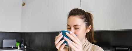 Portrait of carefree smiling woman drinking coffee, standing in kitchen with delighted, pleased face...