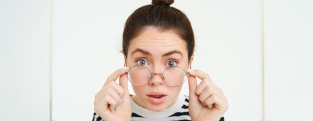 Portrait of woman leaning towards camera as if reading text with surprised face, takes off glasses,...