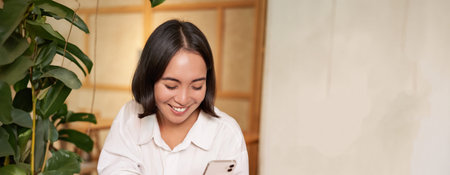 Portrait of modern girl sits with coffee in cafe, smiling while looking at smartphone, reading book...