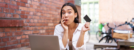 Stylish young asian woman doing shopping online, sitting with credit card and laptop, buying in...