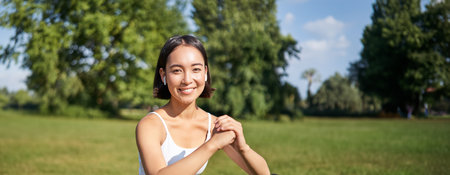 Vertical shot of young fit woman does squats in park, using stretching band on legs, smiling pleased while workoutの写真素材