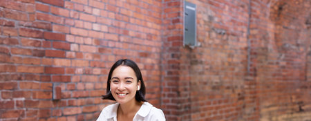 Stylish asian girl works in coworking with laptop, makes notes, uses wireless earphones and smiles,...