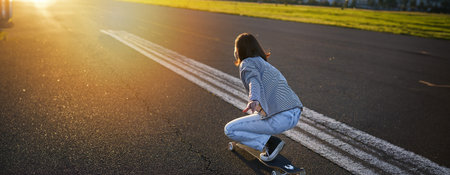 Side view of beautiful asian girl on skateboard, riding her cruiser towards the sun on an empty...