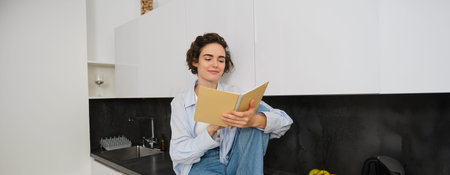 Portrait of modern young woman reading in kitchen, sitting on counter and smiling, studying