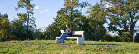 Side view of young woman sitting alone in park on bench, using her laptop to study or work remotely...