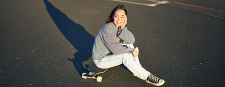Portrait of asian woman sitting on skateboard, skating on her cruiser longboard, using smartphone...