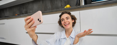 Portrait of young woman video chats, records vlog while sits on kitchen floor. Cute girl takes selfie on smartphoneの写真素材